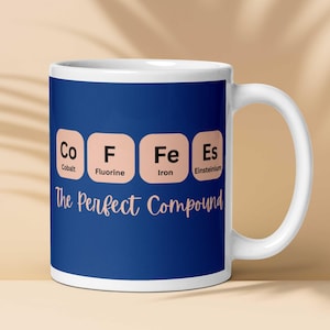 Puede incluir: Taza de cerámica blanca con fondo azul. La taza presenta las palabras "Co F Fe Es" que representan Cobalto, Flúor, Hierro y Einstenio. Debajo, el texto dice "The Perfect Compound".