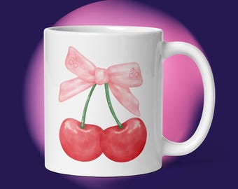 May include: White ceramic mug featuring a watercolor design of two red cherries with green stems and a pink bow. The bow has cherry illustrations. The mug has a white handle and is set against a purple and pink background.