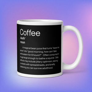 Puede incluir: Taza de café de cerámica blanca con un panel negro con la palabra "Coffee" y una definición humorística. La definición describe los efectos del café, incluyendo convertir "déjame en paz" en "buenos días".