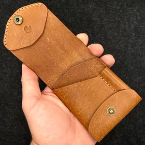 Op de afbeelding: Een bruine leren brillenkoker met een klepsluiting en een messing drukknop. De koker heeft een gestikt ontwerp en een logo op de klep. De koker wordt in een hand gehouden tegen een donkere achtergrond.