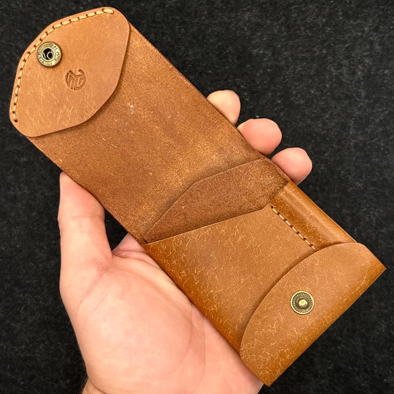 Op de afbeelding: Een bruine leren brillenkoker met een klepsluiting en een messing drukknop. De koker heeft een gestikt ontwerp en een logo op de klep. De koker wordt in een hand gehouden tegen een donkere achtergrond.