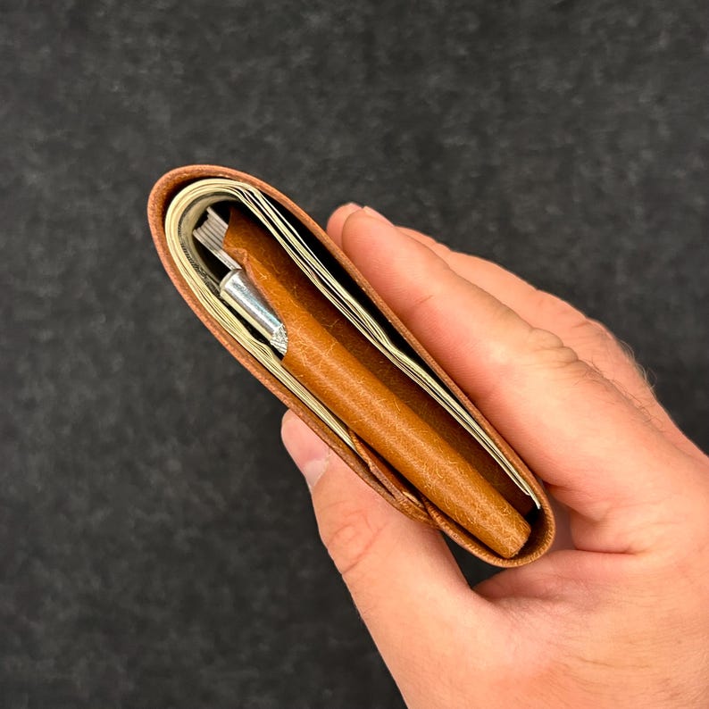 Op de afbeelding: Een bruine leren portemonnee, gedeeltelijk open, met gevouwen bankbiljetten en een zilveren pen. De portemonnee wordt in een hand gehouden tegen een donkere achtergrond. Het leer heeft een textuur.