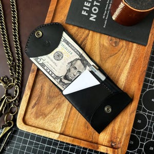 Peut inclure: Un portefeuille en cuir noir avec une fermeture à bouton-pression, contenant un billet de cinq dollars, une petite carte blanche et un stylo argenté. Le portefeuille est sur un plateau en bois, avec un carnet et un objet en cuir en arrière-plan.
