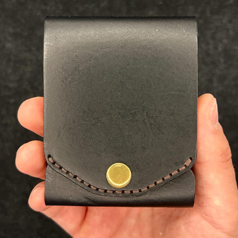Op de afbeelding: Een zwarte leren portemonnee met een messing knoopsluiting. De rechthoekige portemonnee heeft afgeronde randen en bruine stiksels op de flap. Het item wordt in een hand gehouden.