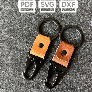 May include: Two keyrings with black metal clasps and rings. Each keyring has a tan leather strap with a black rivet. The background is a textured grey surface. Above the keyrings are white icons with the letters PDF, SVG, and DXF.