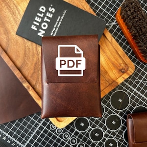 Könnte beinhalten: Ein brauner Lederkartenhalter mit weißem PDF-Symbol. Ein schwarzes Notizbuch mit "FIELD NOTES"-Aufdruck ist sichtbar. Der Hintergrund umfasst ein Holzschneidebrett und eine Bürste.