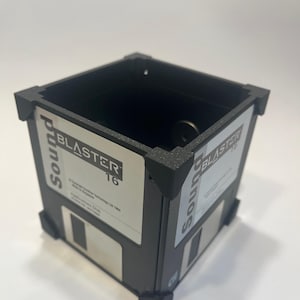 Puede incluir: Un macetero negro en forma de cubo hecho de disquetes reciclados. El macetero tiene el texto "Sound Blaster 16" en un lado y "Sound Blaster Pro" en el otro.