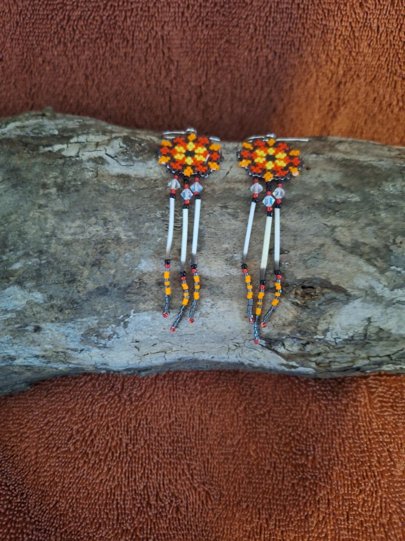 May include: A pair of beaded earrings with a geometric pattern in orange, yellow, and black. The earrings have a crystal bead and a white feather dangle.