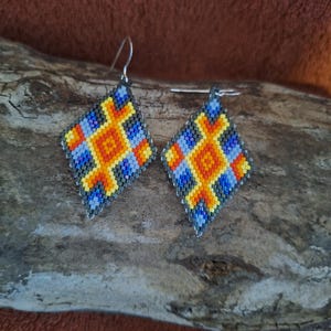 May include: A pair of beaded earrings with a geometric design in shades of blue, yellow, orange, and black. The earrings are diamond-shaped and have silver hooks.