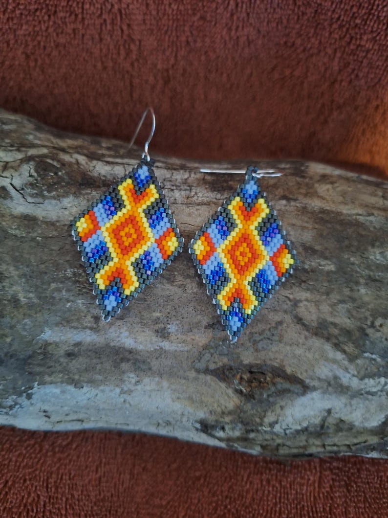 May include: A pair of beaded earrings with a geometric design in shades of blue, yellow, orange, and black. The earrings are diamond-shaped and have silver hooks.