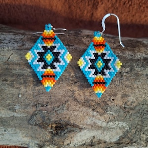 May include: A pair of diamond-shaped beaded earrings featuring a geometric pattern in turquoise, black, white, yellow, and orange. The earrings are constructed from small glass beads and include silver-toned metal hooks for attachment to the ears.