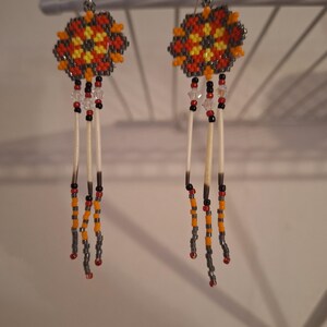 May include: A pair of beaded earrings with a geometric design in shades of orange, yellow, and gray. The earrings feature a long, dangling design with white feathers and beads.
