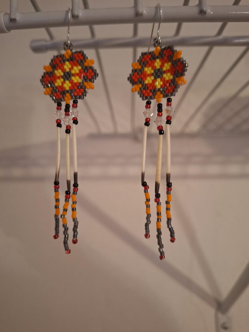 May include: A pair of beaded earrings with a geometric design in shades of orange, yellow, and gray. The earrings feature a long, dangling design with white feathers and beads.