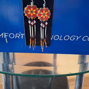 May include: A pair of beaded earrings with a geometric design in red, orange, and yellow. The earrings are hanging from silver hooks and have a fringe of beads.