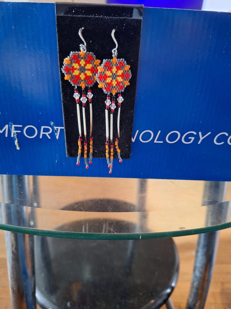 May include: A pair of beaded earrings with a geometric design in red, orange, and yellow. The earrings are hanging from silver hooks and have a fringe of beads.