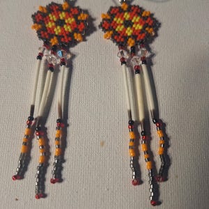 May include: A pair of beaded earrings with a geometric design in shades of red, orange, yellow, black, and white. The earrings feature a long dangle with white feathers and beads.