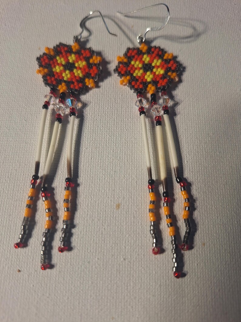 May include: A pair of beaded earrings with a geometric design in shades of red, orange, yellow, black, and white. The earrings feature a long dangle with white feathers and beads.