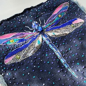 May include: A close-up of a blue, purple, and green beaded dragonfly embroidered on a dark blue fabric. The dragonfly has a large, iridescent blue gem in its center.