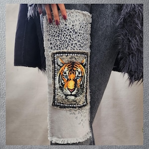 May include: A pair of gray denim jeans with a white patch featuring a tiger face. The patch is decorated with rhinestones and has a leopard print background.