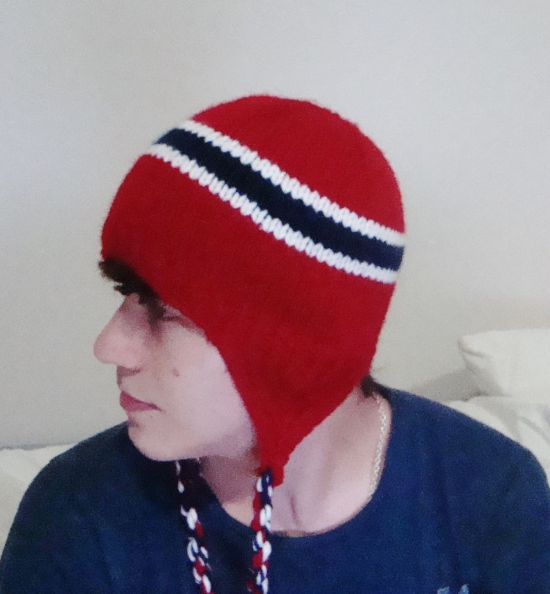 May include: A red knit hat with a white and blue striped band. The hat has ear flaps that tie under the chin.