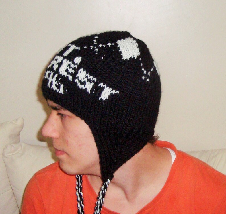May include: Black knit hat with ear flaps and white text. The hat has a geometric pattern and the text "FIRE" is visible. The hat has braided black and white ties.