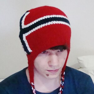 May include: A red knitted hat with white and black stripes, resembling a Scandinavian flag. The hat has a long, dangling tassel.