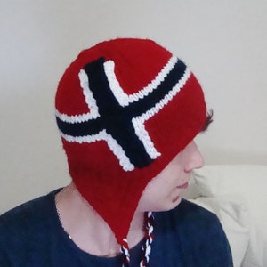 May include: A red knitted hat with a white and black Norwegian flag design. The hat has ear flaps and a long, braided cord.