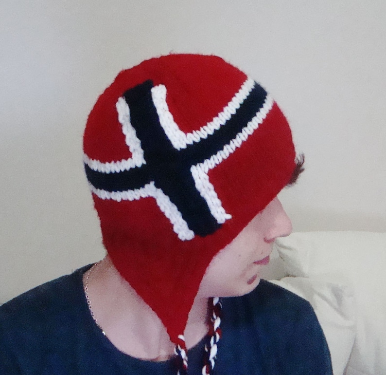 May include: A red knitted hat with a white and black Norwegian flag design. The hat has ear flaps and a long, braided cord.