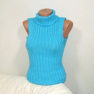May include: A sleeveless, turquoise blue turtleneck sweater displayed on a mannequin. The ribbed knit top has a high, folded collar and a fitted silhouette. The sweater is made of a soft, possibly wool or cotton blend.