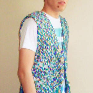 May include: A man wearing a white t-shirt with a blue and green crocheted vest. The vest has a fringe trim and is made of a variety of colors.