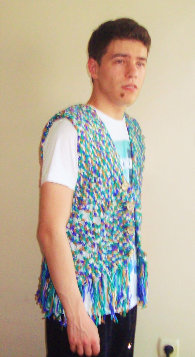 May include: A man wearing a white t-shirt with a blue and green crocheted vest. The vest has a fringe trim and is made of a variety of colors.