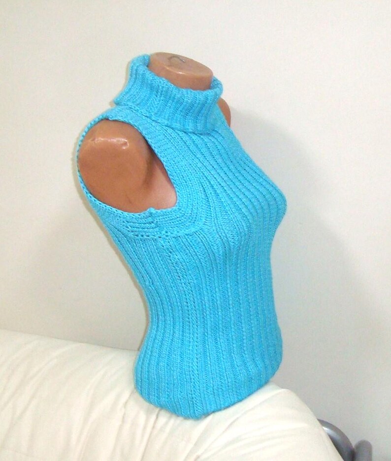 May include: A sleeveless, turquoise blue knitted turtleneck top. The top is displayed on a mannequin, showcasing its ribbed texture and form-fitting design. The garment features a high, folded turtleneck and a scoop neckline.