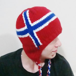May include: A red knitted hat with a white and blue Norwegian flag design. The hat has ear flaps and a long tassel.