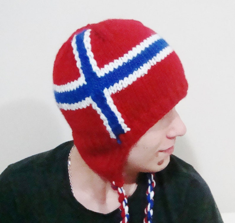 May include: A red knitted hat with a white and blue Norwegian flag design. The hat has ear flaps and a long tassel.