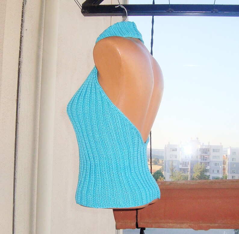 May include: A turquoise halter top made of knitted fabric. The top features a ribbed texture and a high neckline that transitions into a halter design. The back is open, and the top is displayed on a mannequin.