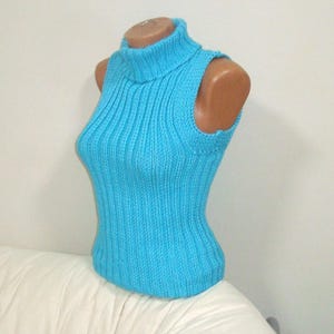 May include: A sleeveless, turquoise blue turtleneck sweater displayed on a mannequin. The sweater features a ribbed knit design and a high neckline. The garment is made of a soft, possibly wool or cotton, material.
