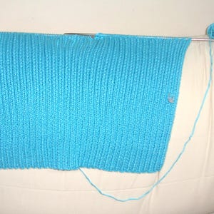May include: A partially completed knitted project in a vibrant turquoise blue. The knitting is on metal needles, with several stitch markers in place. A ball of matching yarn sits nearby, with the yarn extending to the project.