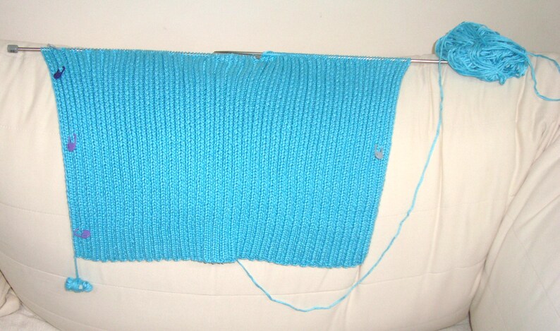 May include: A partially completed knitted project in a vibrant turquoise blue. The knitting is on metal needles, with several stitch markers in place. A ball of matching yarn sits nearby, with the yarn extending to the project.