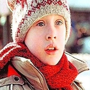 May include: A person wearing a red and white patterned knitted hat and a red scarf. They are also wearing a tan jacket with a zip. The image is a medium shot, focusing on the person's face and upper body.