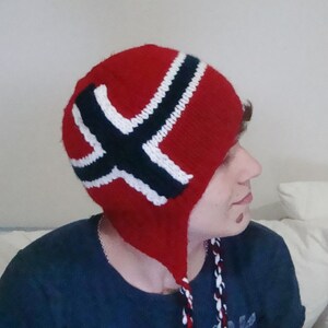 May include: A red knitted hat with a white and black Norwegian flag design. The hat has ear flaps and a long tassel.