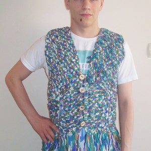 May include: A person wearing a multi-colored crocheted vest with fringe. The vest has brown, blue, green, and white yarn. The vest has a V-neck and buttons down the front.