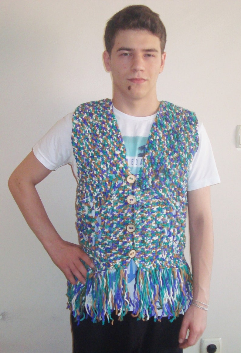 May include: A person wearing a multi-colored crocheted vest with fringe. The vest has brown, blue, green, and white yarn. The vest has a V-neck and buttons down the front.