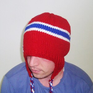 May include: A red knitted hat with white and blue stripes. The hat has ear flaps that are tied together at the chin.