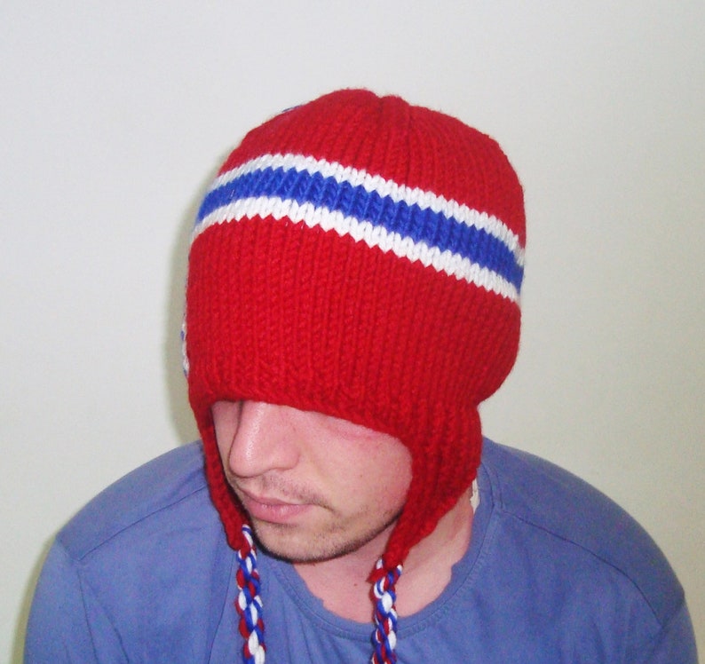 May include: A red knitted hat with white and blue stripes. The hat has ear flaps that are tied together at the chin.