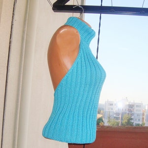 May include: A turquoise blue knitted halter top with a ribbed texture. The top features a high neck and an open back design, displayed on a mannequin. The garment is hanging on a hanger against a bright outdoor background.