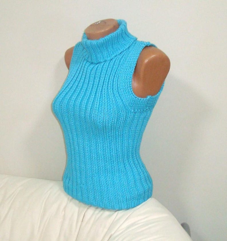 May include: A sleeveless, turquoise blue turtleneck sweater displayed on a mannequin. The ribbed knit top has a fitted silhouette and a high neckline. The garment is made of a soft, textured material, and is suitable for layering or wearing on its own.