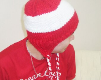 May include: A red and white knit hat with ear flaps and braided tassels. The hat features a horizontal white stripe across the top. The person in the image is also wearing a red t-shirt with the text "Ocean Club".
