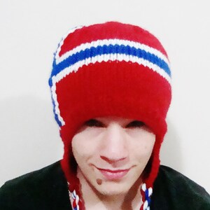 May include: A red knit hat with white and blue stripes, resembling a national flag. The hat has ear flaps and a pompom on top.