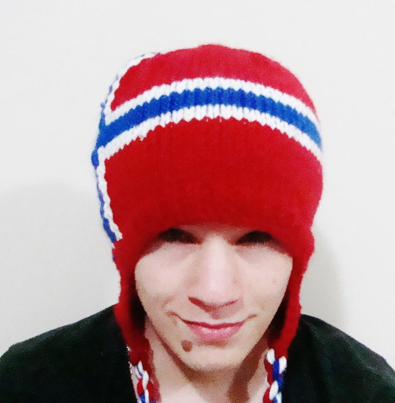 May include: A red knit hat with white and blue stripes, resembling a national flag. The hat has ear flaps and a pompom on top.