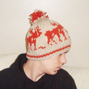 May include: A beige knitted hat featuring a red reindeer design and a red pom-pom. The hat has a red band at the bottom. This winter hat is made of a soft, warm material, ideal for cold weather.